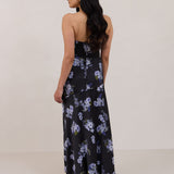 A woman is facing the back, wearing a dark blue strapless dress with a light blue floral print. The dress is fitted in the bodice and hip then flows into a fuller skirt. The dress is tea-length. This is the Sterling Dress in Viola.