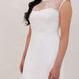 A woman stands against a white backdrop. She's wearing a delicate lace overlay dress on top of a simple white strapless mini bridal dress. This is the Winslow Overlay.
