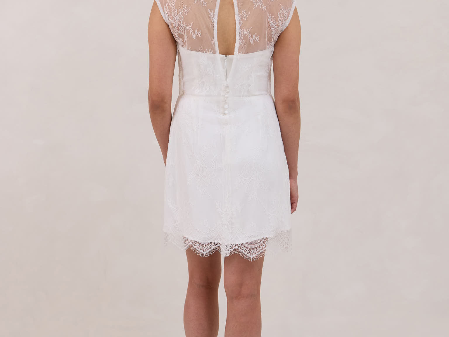 A woman stands against a white backdrop. She's wearing a delicate lace overlay dress on top of a simple white strapless mini bridal dress. This is the Winslow Overlay.