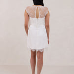 A woman stands against a white backdrop. She's wearing a delicate lace overlay dress on top of a simple white strapless mini bridal dress. This is the Winslow Overlay.