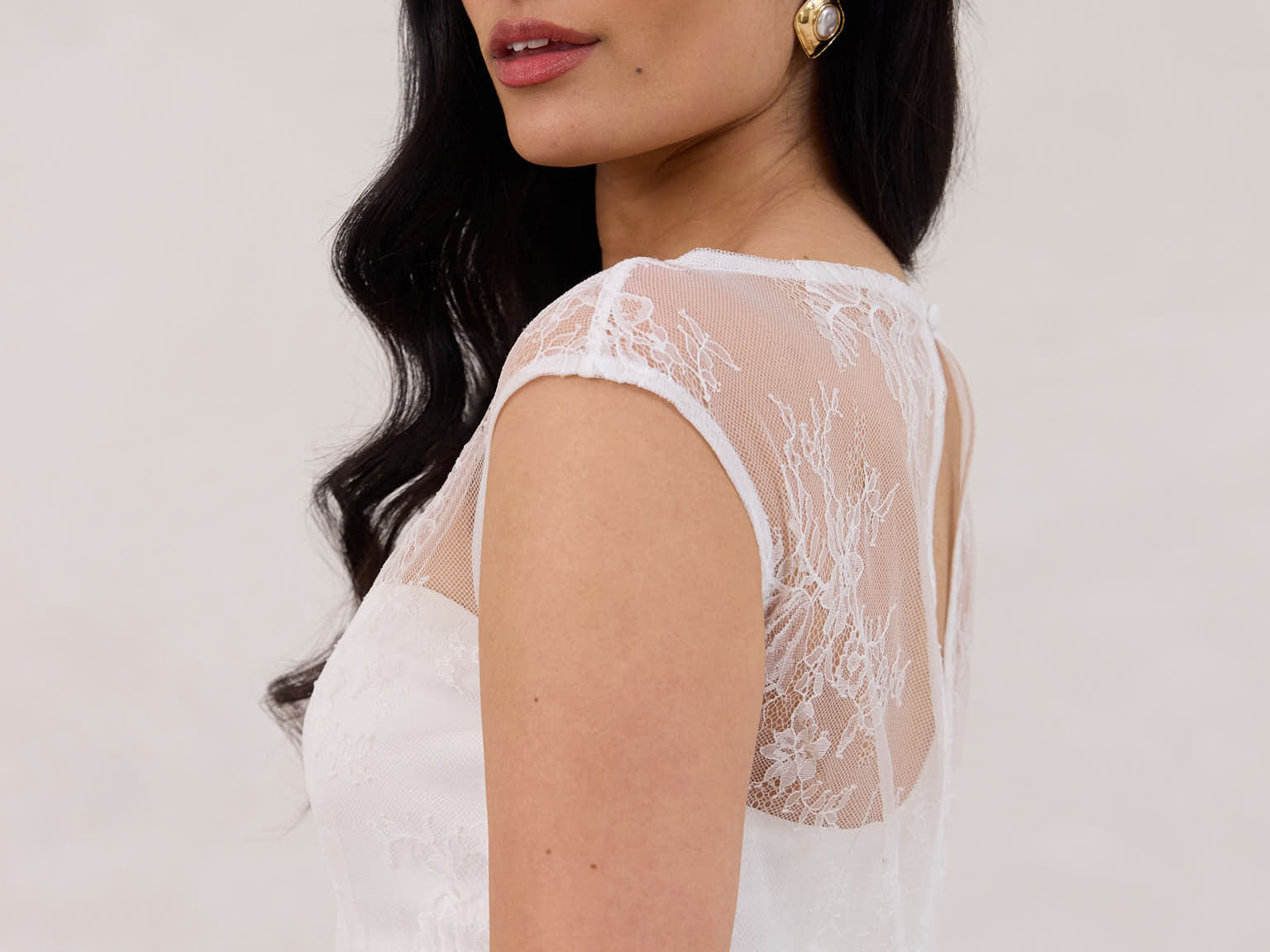 A woman stands against a white backdrop. She's wearing a delicate lace overlay dress on top of a simple white strapless mini bridal dress. This is the Winslow Overlay.