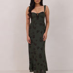 A woman is standing infront of a floral display. She's wearing a dark green special occasion dress with a dark brown floral print. The dress s fitted in the bodice with a little keyhole detail and tie at the bust. The dress is tea-length and has shirring at the back for added comfort. This is the Decker dress in Esme.