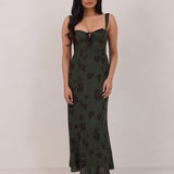 A woman is standing infront of a floral display. She's wearing a dark green special occasion dress with a dark brown floral print. The dress s fitted in the bodice with a little keyhole detail and tie at the bust. The dress is tea-length and has shirring at the back for added comfort. This is the Decker dress in Esme.