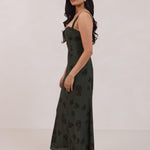 A woman is standing infront of a floral display. She's wearing a dark green special occasion dress with a dark brown floral print. The dress s fitted in the bodice with a little keyhole detail and tie at the bust. The dress is tea-length and has shirring at the back for added comfort. This is the Decker dress in Esme.