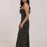 A woman is standing infront of a floral display. She's wearing a dark green special occasion dress with a dark brown floral print. The dress s fitted in the bodice with a little keyhole detail and tie at the bust. The dress is tea-length and has shirring at the back for added comfort. This is the Decker dress in Esme.
