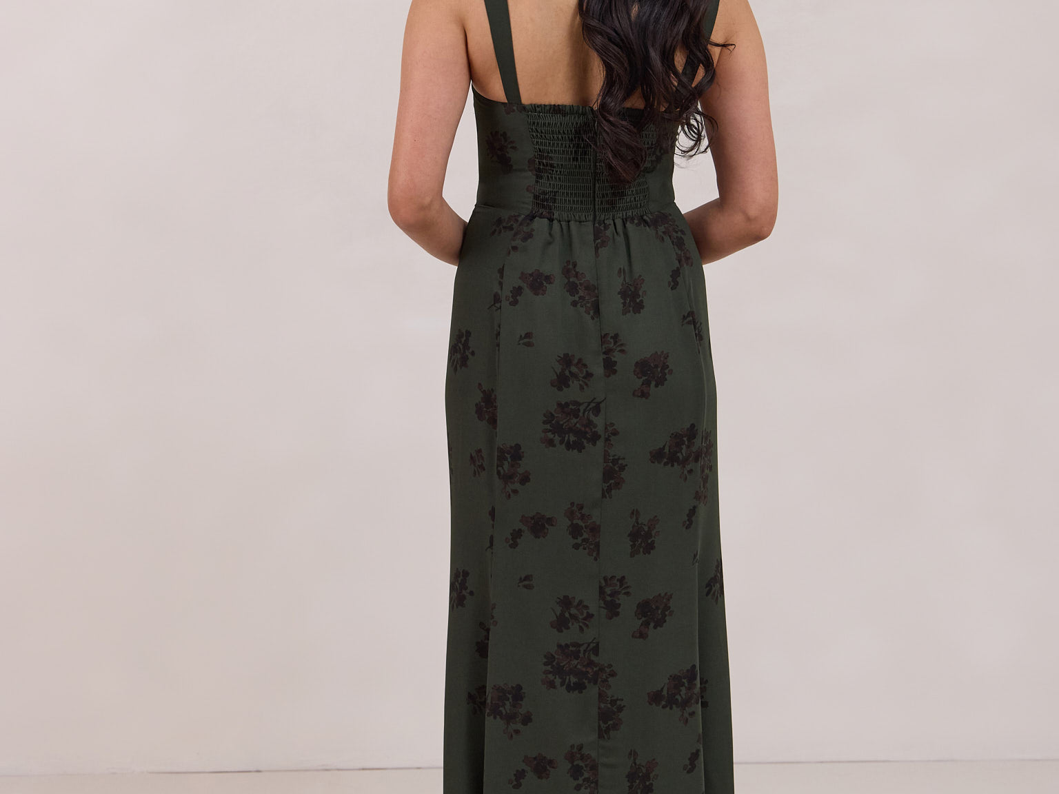 A woman is standing infront of a floral display. She's wearing a dark green special occasion dress with a dark brown floral print. The dress s fitted in the bodice with a little keyhole detail and tie at the bust. The dress is tea-length and has shirring at the back for added comfort. This is the Decker dress in Esme.
