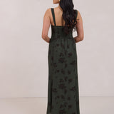 A woman is standing infront of a floral display. She's wearing a dark green special occasion dress with a dark brown floral print. The dress s fitted in the bodice with a little keyhole detail and tie at the bust. The dress is tea-length and has shirring at the back for added comfort. This is the Decker dress in Esme.