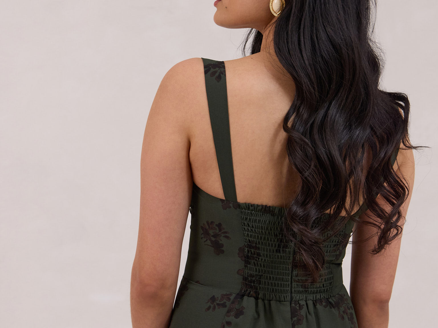 A woman is standing infront of a floral display. She's wearing a dark green special occasion dress with a dark brown floral print. The dress s fitted in the bodice with a little keyhole detail and tie at the bust. The dress is tea-length and has shirring at the back for added comfort. This is the Decker dress in Esme.