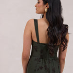 A woman is standing infront of a floral display. She's wearing a dark green special occasion dress with a dark brown floral print. The dress s fitted in the bodice with a little keyhole detail and tie at the bust. The dress is tea-length and has shirring at the back for added comfort. This is the Decker dress in Esme.