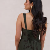 A woman is standing infront of a floral display. She's wearing a dark green special occasion dress with a dark brown floral print. The dress s fitted in the bodice with a little keyhole detail and tie at the bust. The dress is tea-length and has shirring at the back for added comfort. This is the Decker dress in Esme.