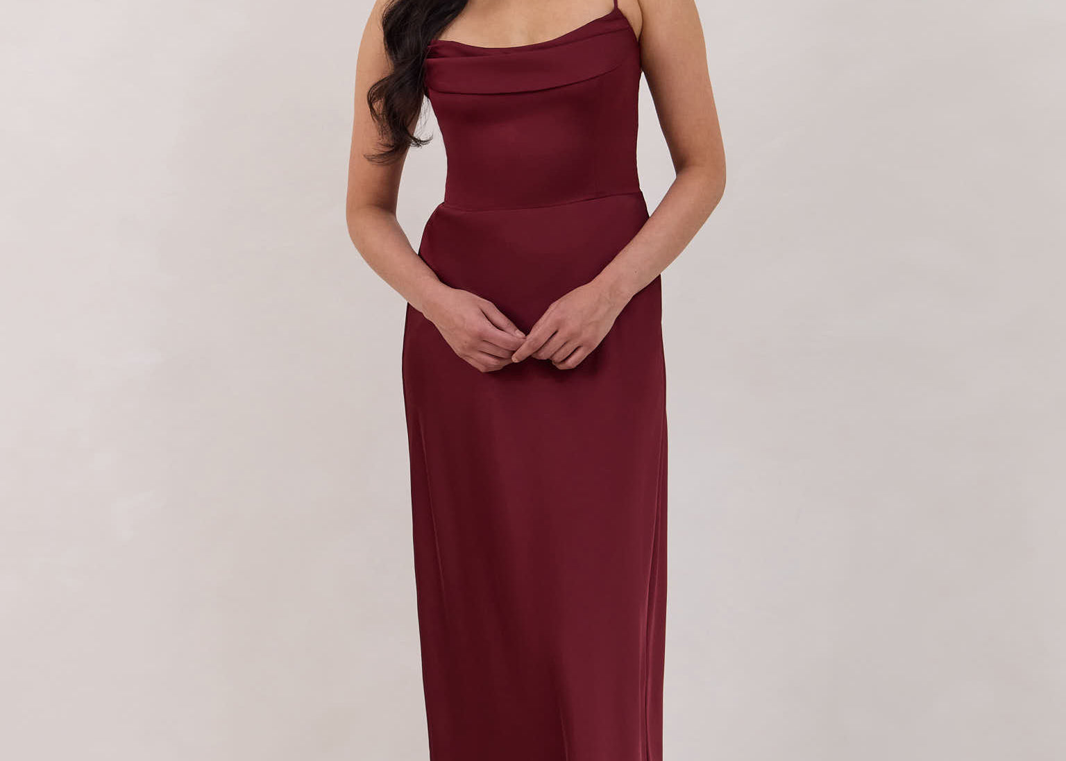 A woman is standing against a white backdrop wearing a burgundy, tea length satin bridesmaid dress. The dress has a boned bodice with a slight gathering at the bust, and a bias cut skirt that falls nicely over the hips. This is the Rosedale dress in merlot.