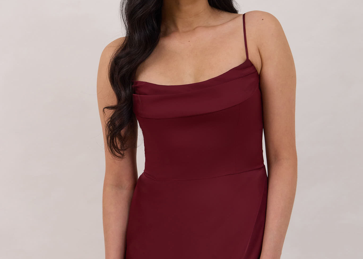 A woman is standing against a white backdrop wearing a burgundy, tea length satin bridesmaid dress. The dress has a boned bodice with a slight gathering at the bust, and a bias cut skirt that falls nicely over the hips. This is the Rosedale dress in merlot.