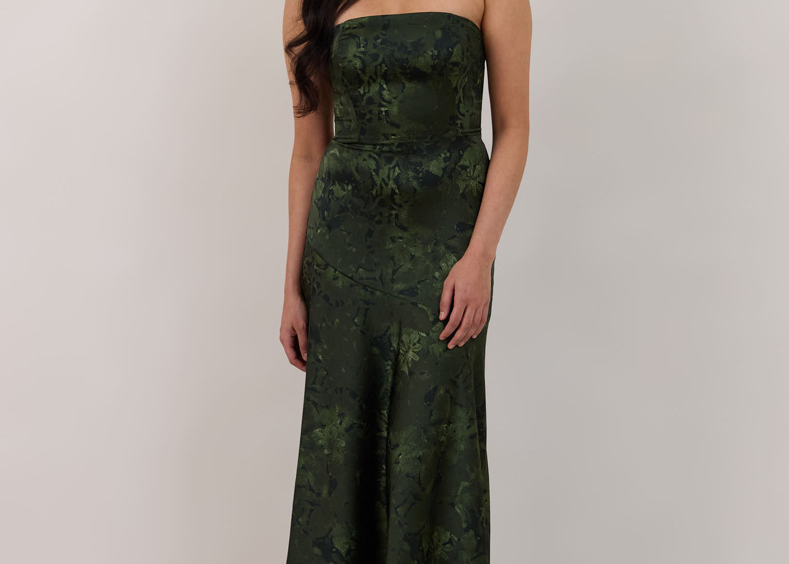 A woman standing against a white backdrop wearing a strapless tea length dress. The dress is a dark green with a subtle green floral print. The dress is fitted through the bodice, and has a more flowy skirt. This is the Sterling dress in Fern.