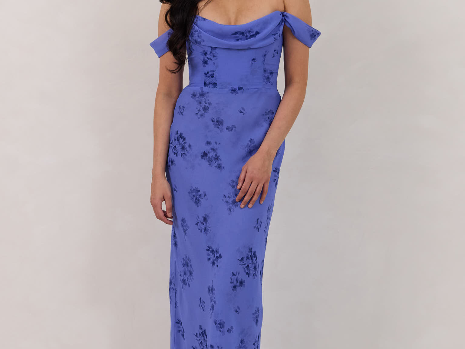 A woman standing against a white background wearing a periwinkle blue floral print dress. The dress has a scooped neckline with an off-the-shoulder strap, is fitted through the bodice and falls to ankle length. The fabric is chiffon. This is the Sutton Dress in Livia.