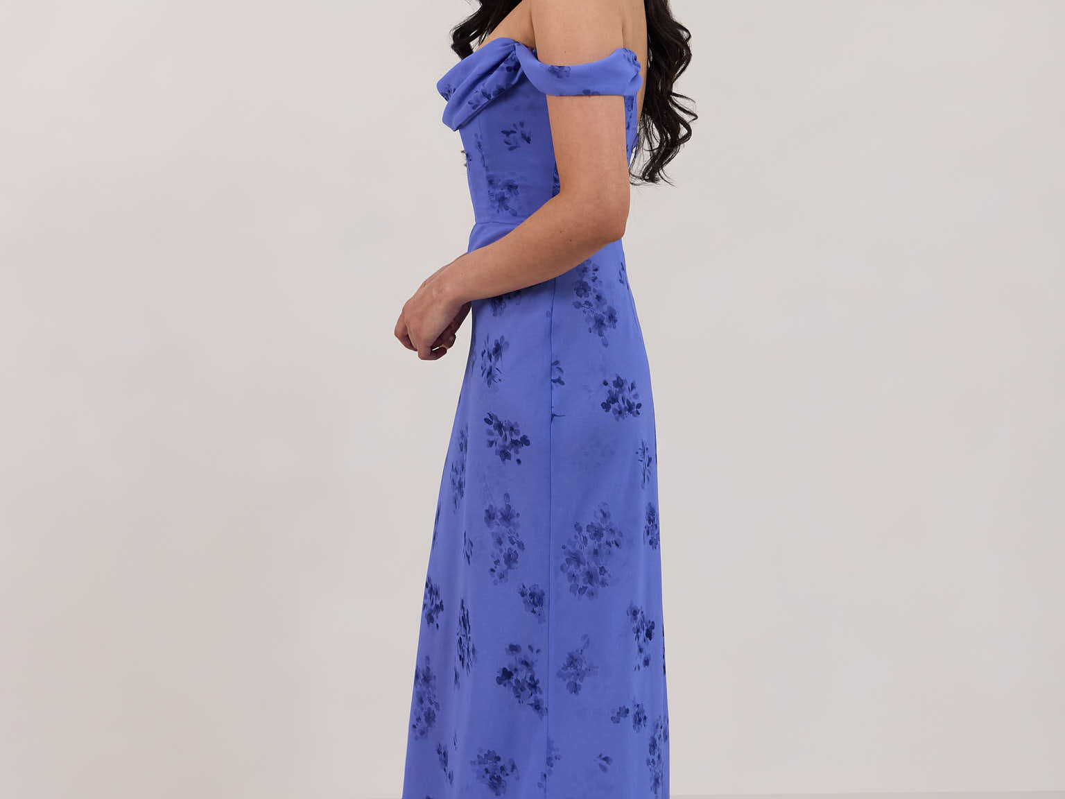 A woman standing facing the left against a white background wearing a periwinkle blue floral print dress. The dress has a scooped neckline with an off-the-shoulder strap, is fitted through the bodice and falls to ankle length. The fabric is chiffon. This is the Sutton Dress in Livia.