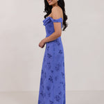 A woman standing facing the left against a white background wearing a periwinkle blue floral print dress. The dress has a scooped neckline with an off-the-shoulder strap, is fitted through the bodice and falls to ankle length. The fabric is chiffon. This is the Sutton Dress in Livia.