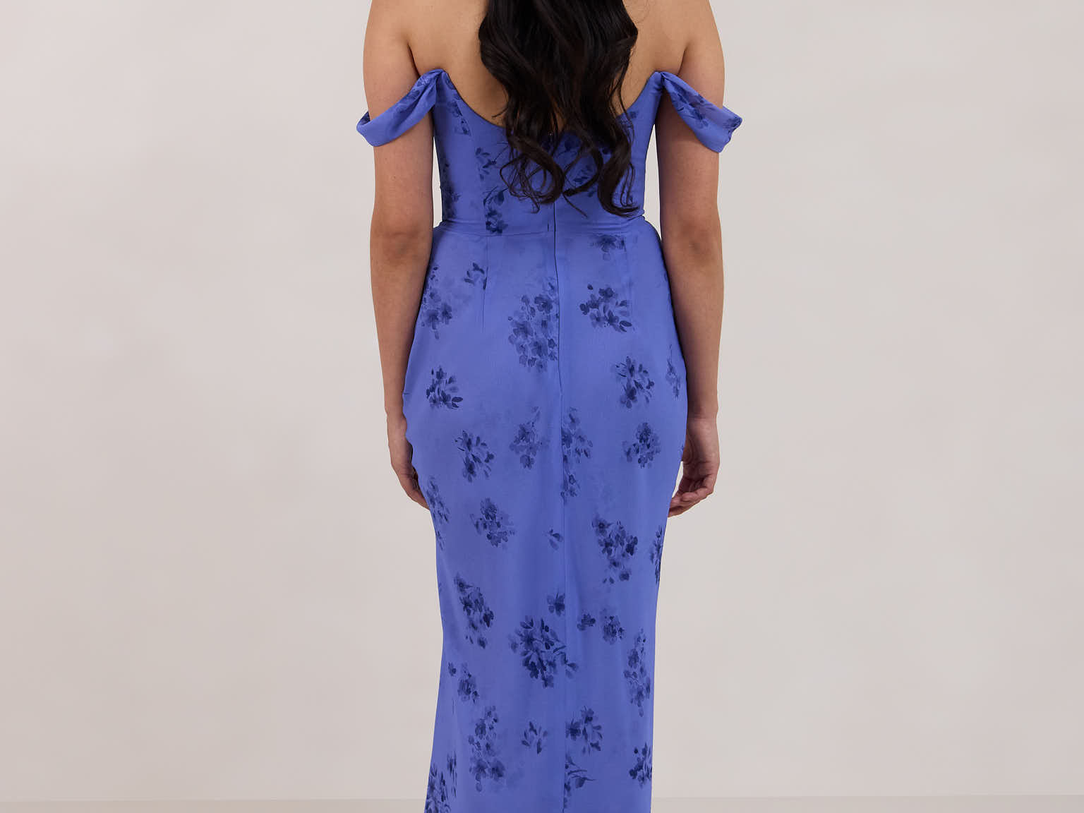 A woman standing facing the back against a white background wearing a periwinkle blue floral print dress. The dress has a scooped neckline with an off-the-shoulder strap, is fitted through the bodice and falls to ankle length. The fabric is chiffon. This is the Sutton Dress in Livia.