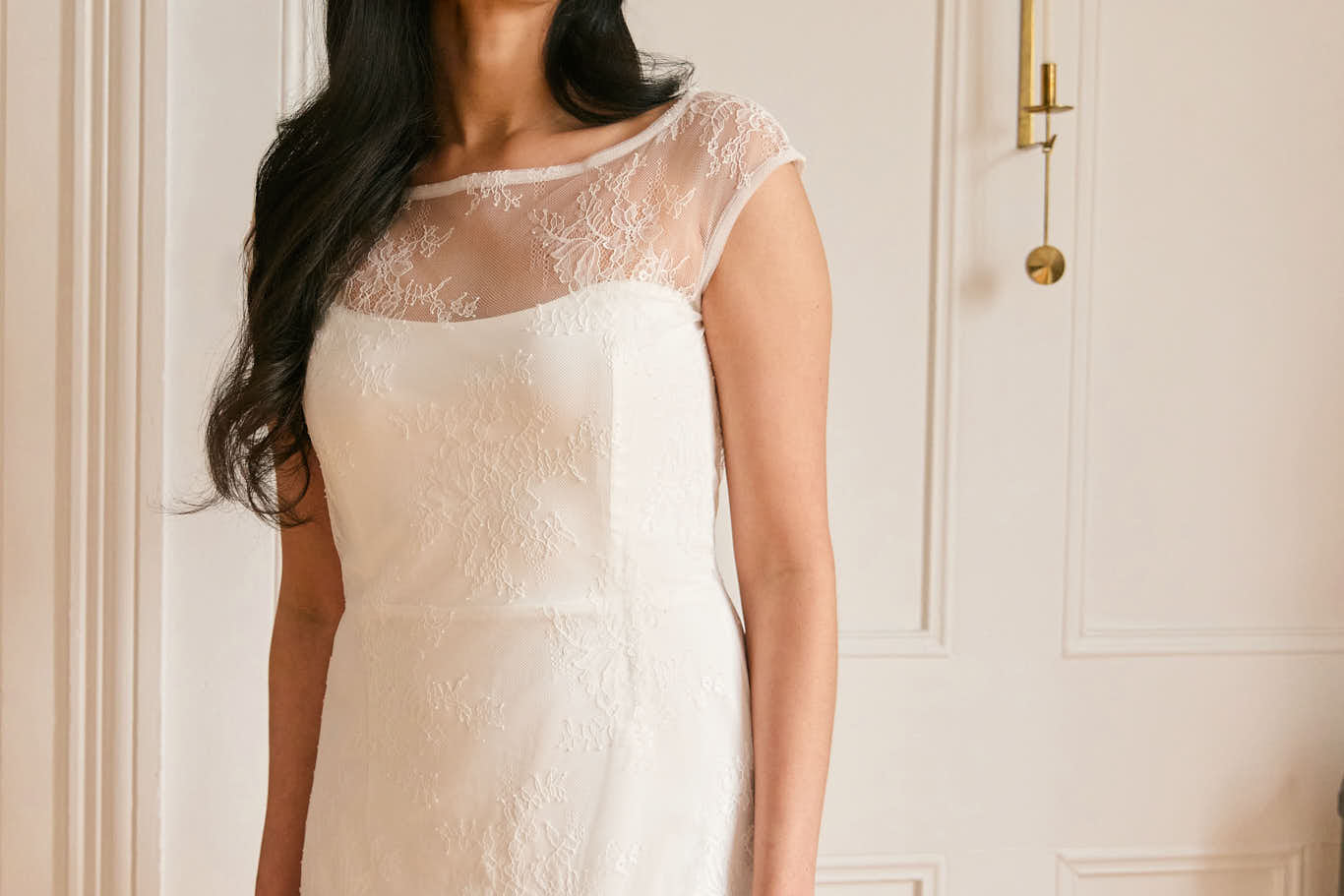 A woman stands against a white backdrop. She's wearing a delicate lace overlay dress on top of a simple white strapless mini bridal dress. This is the Winslow Overlay.