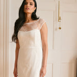 A woman stands in a white apartment. She's wearing a delicate lace overlay dress on top of a simple white strapless mini bridal dress. This is the Winslow Overlay.