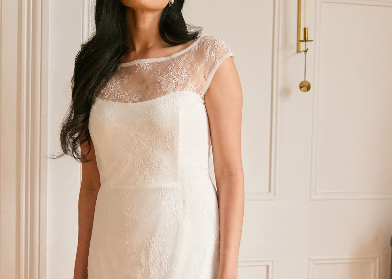 A woman stands in a white apartment. She's wearing a delicate lace overlay dress on top of a simple white strapless mini bridal dress. This is the Winslow Overlay.