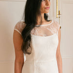 A woman stands against a white backdrop. She's wearing a delicate lace overlay dress on top of a simple white strapless mini bridal dress. This is the Winslow Overlay.