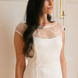 A woman stands against a white backdrop. She's wearing a delicate lace overlay dress on top of a simple white strapless mini bridal dress. This is the Winslow Overlay.