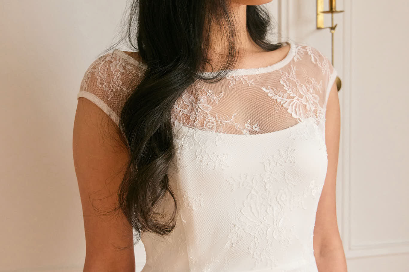 A woman stands against a white backdrop. She's wearing a delicate lace overlay dress on top of a simple white strapless mini bridal dress. This is the Winslow Overlay.