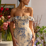 A woman is standing in front of floral arrangements. She's wearing a tea-length cream dress with a delicate blue floral print. The dress has a ruffle detail around the neckline that can be worn on or off the shoulder. The skirt has a slit up to the thigh and a ruffle detail around the hem. This is the Renfrew Dress in Ayla.