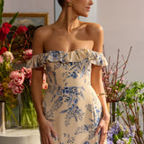 A woman is standing in front of floral arrangements. She's wearing a tea-length cream dress with a delicate blue floral print. The dress has a ruffle detail around the neckline that can be worn on or off the shoulder. The skirt has a slit up to the thigh and a ruffle detail around the hem. This is the Renfrew Dress in Ayla.