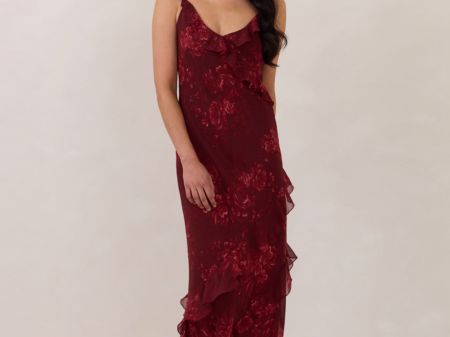 Model wearing the Majorca Dress in Garnet. A full length flowy guesting dress with a deep v neckline and ruffle details. Garnet is a dark red floral print.