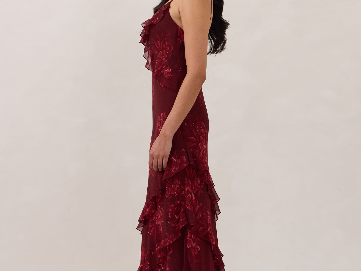 Side shot of model wearing the Majorca Dress in Garnet. A full length flowy guesting dress with a deep v neckline and ruffle details. Garnet is a dark red floral print.