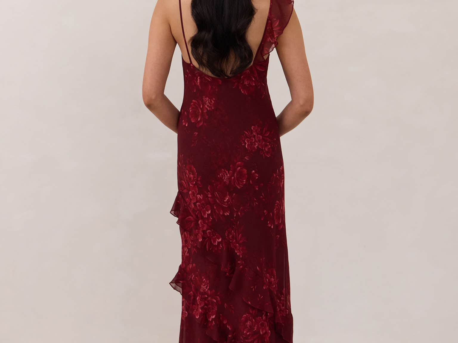 Back shot of model wearing the Majorca Dress in Garnet. A full length flowy guesting dress with a deep v neckline and ruffle details. Garnet is a dark red floral print.