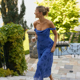 A woman walking up outdoor patio stairs holding a glass of champagne. She's wearing a periwinkle blue chiffon dress with a subtle dark blue floral print. The dress has a scoop neckline with an off the shoulder detail.