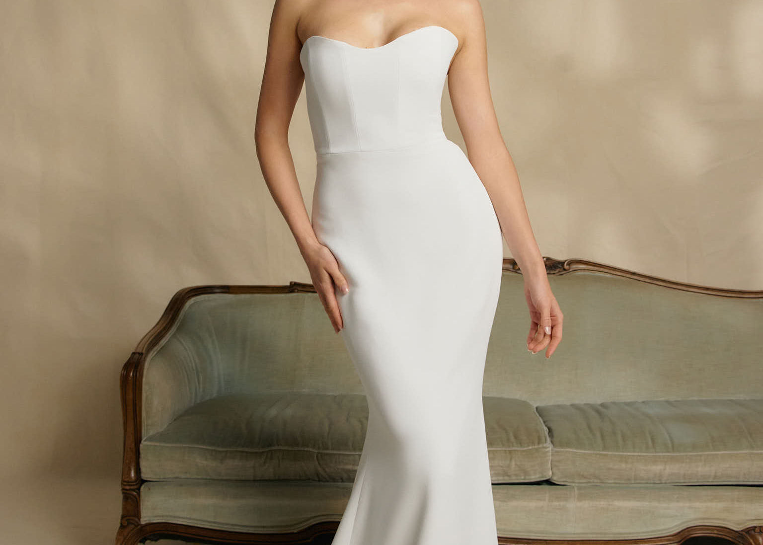 Woman in a strapless white wedding dress standing in front of a vintage couch.