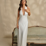 Woman in a white halter bridal dress standing in front of a vintage couch against a beige wall.