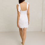A woman stands against a light grey backdrop. She's wearing a white mini dress with white floral embroidery and satin tie straps. This bridal event dress is called the Tanner Mini.