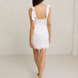 A woman stands against a light grey backdrop. She's wearing a white mini dress with white floral embroidery and satin tie straps. This bridal event dress is called the Tanner Mini.