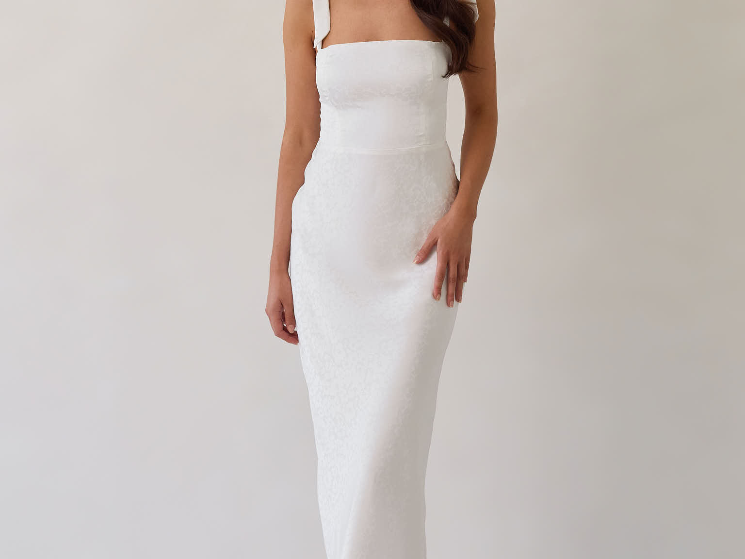 A woman is wearing a white floral jacquard dress with tie straps. The dress has a square neckline, is fitted through the bodice and then skims the hips nicely with a slit in the back. The dress is tea length. This is the Dalton Dress in White Jacquard.