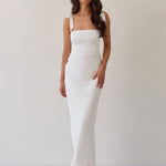 A woman is wearing a white floral jacquard dress with tie straps. The dress has a square neckline, is fitted through the bodice and then skims the hips nicely with a slit in the back. The dress is tea length. This is the Dalton Dress in White Jacquard.
