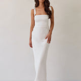 A woman is wearing a white floral jacquard dress with tie straps. The dress has a square neckline, is fitted through the bodice and then skims the hips nicely with a slit in the back. The dress is tea length. This is the Dalton Dress in White Jacquard.