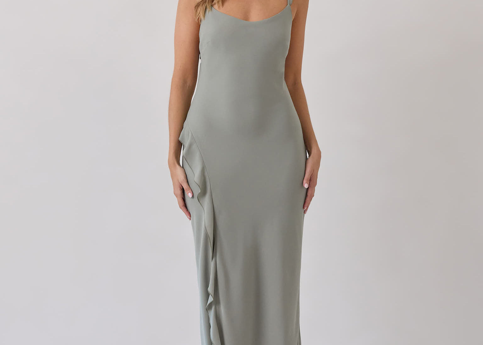 A woman is standing against a white backdrop wearing a sage green bridesmaid dress with an open back. The dress features a slight ruffle detail along the strap and slit of the skirt. This is the Avondale Dress in True Sage. 