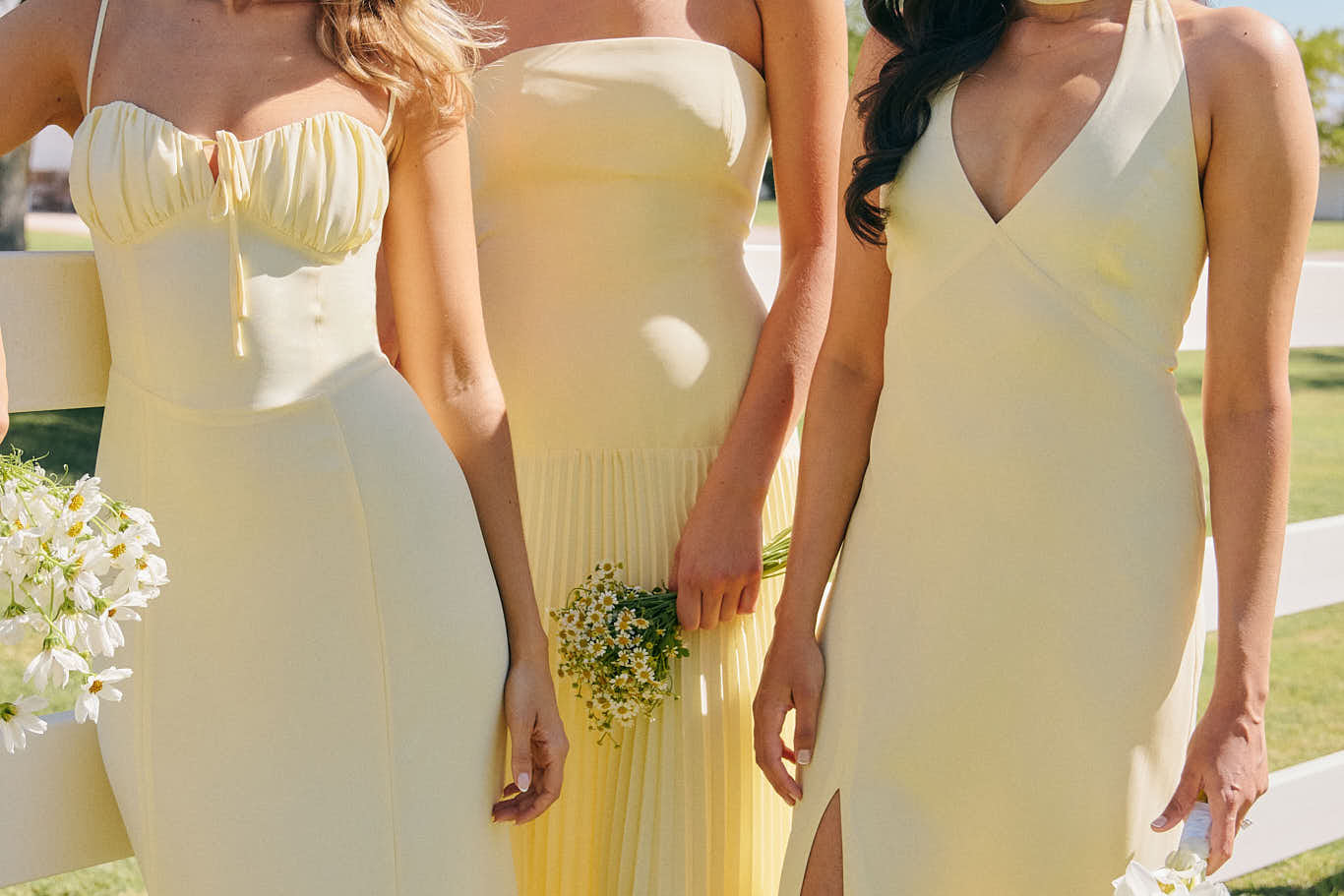 Three woman are standing against a fence wearing butter yellow, full length bridesmaid dresses in various styles.