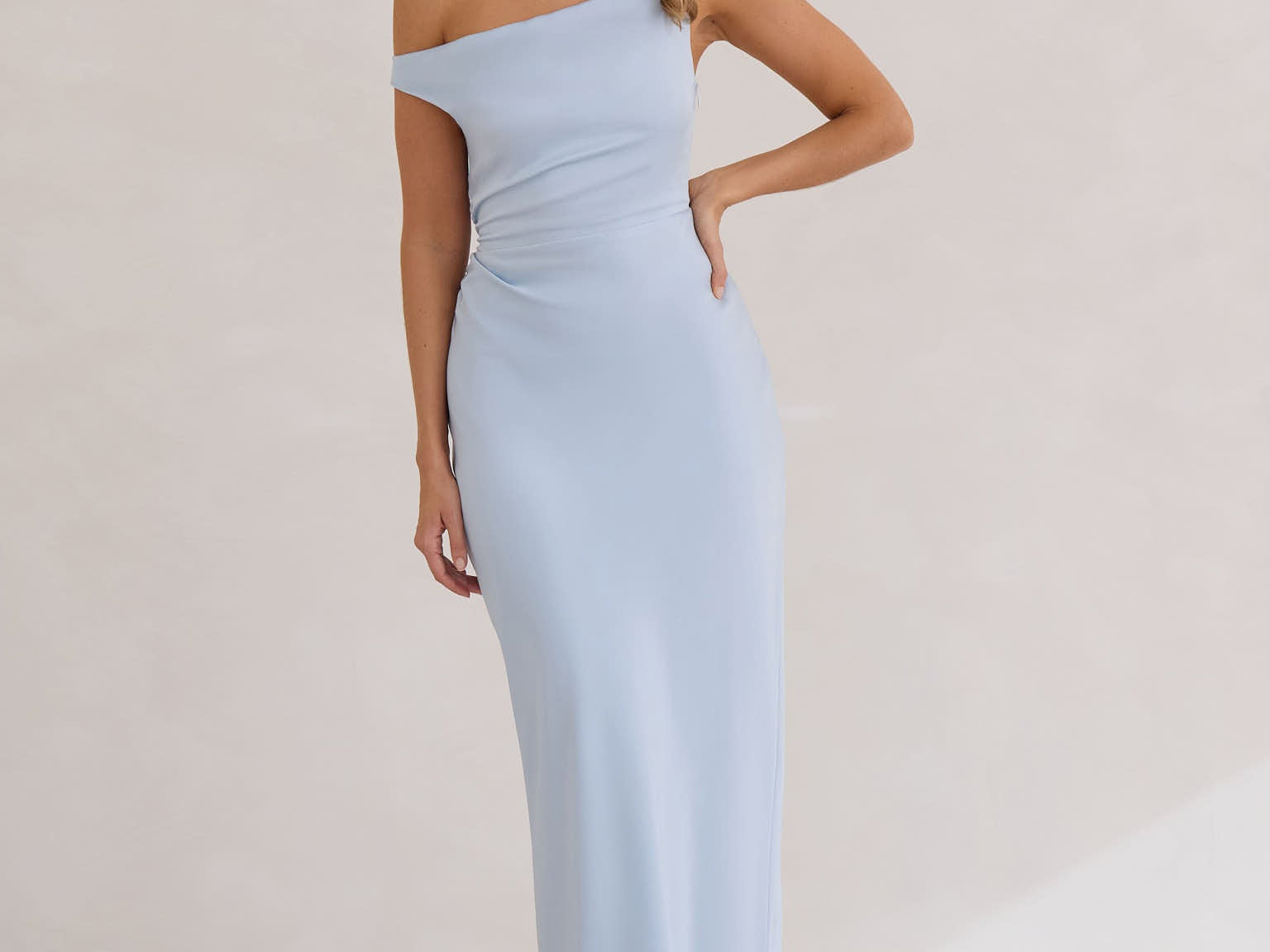 A woman stands infront of a white back drop wearing a powder blue bridesmaid dress. This dress has an asymmetrical neckline and gathers slightly at the waist. Perfect as a bridesmaid dress or a special event dress. This is the Savannah Dress in Powder Blue.