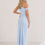 A woman stands infront of a white back drop facing the right wearing a powder blue bridesmaid dress. This dress has an asymmetrical neckline and gathers slightly at the waist. Perfect as a bridesmaid dress or a special event dress. This is the Savannah Dress in Powder Blue.