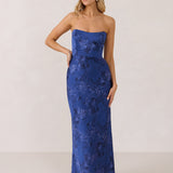A woman stands against a white backdrop. She's wearing an indigo blue strapless dress with a blue floral print. The dress is fitted through the bodice, and falls nicely over the hips and is tea-length. This is the Brodie Dress in Azure. 
