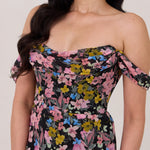 A woman is standing in front of a white background. She's wearing a bold pink and blue floral print strapless dress with an off-the-shoulder strap. This dress is Sutton in Liora and is perfect as a wedding guest dress, or for any special event. 