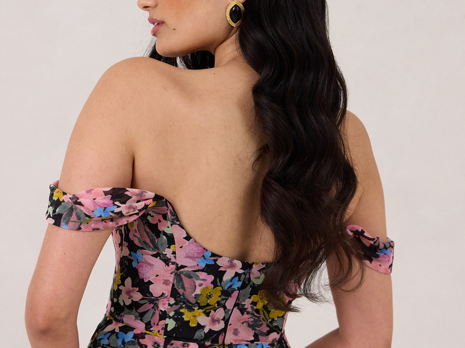 A woman is standing in front of a white background facing the back. She's wearing a bold pink and blue floral print strapless dress with an off-the-shoulder strap. This dress is Sutton in Liora and is perfect as a wedding guest dress, or for any special event. 