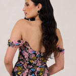 A woman is standing in front of a white background facing the back. She's wearing a bold pink and blue floral print strapless dress with an off-the-shoulder strap. This dress is Sutton in Liora and is perfect as a wedding guest dress, or for any special event. 