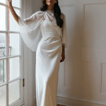 A woman is standing against a window. She's wearing a white bridal dress with a chiffon cape over her shoulders. The cape is gathered slightly on the left side and is nearly fingertip length. This is the Cleo Cape.