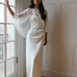 A woman is standing against a window. She's wearing a white bridal dress with a chiffon cape over her shoulders. The cape is gathered slightly on the left side and is nearly fingertip length. This is the Cleo Cape.